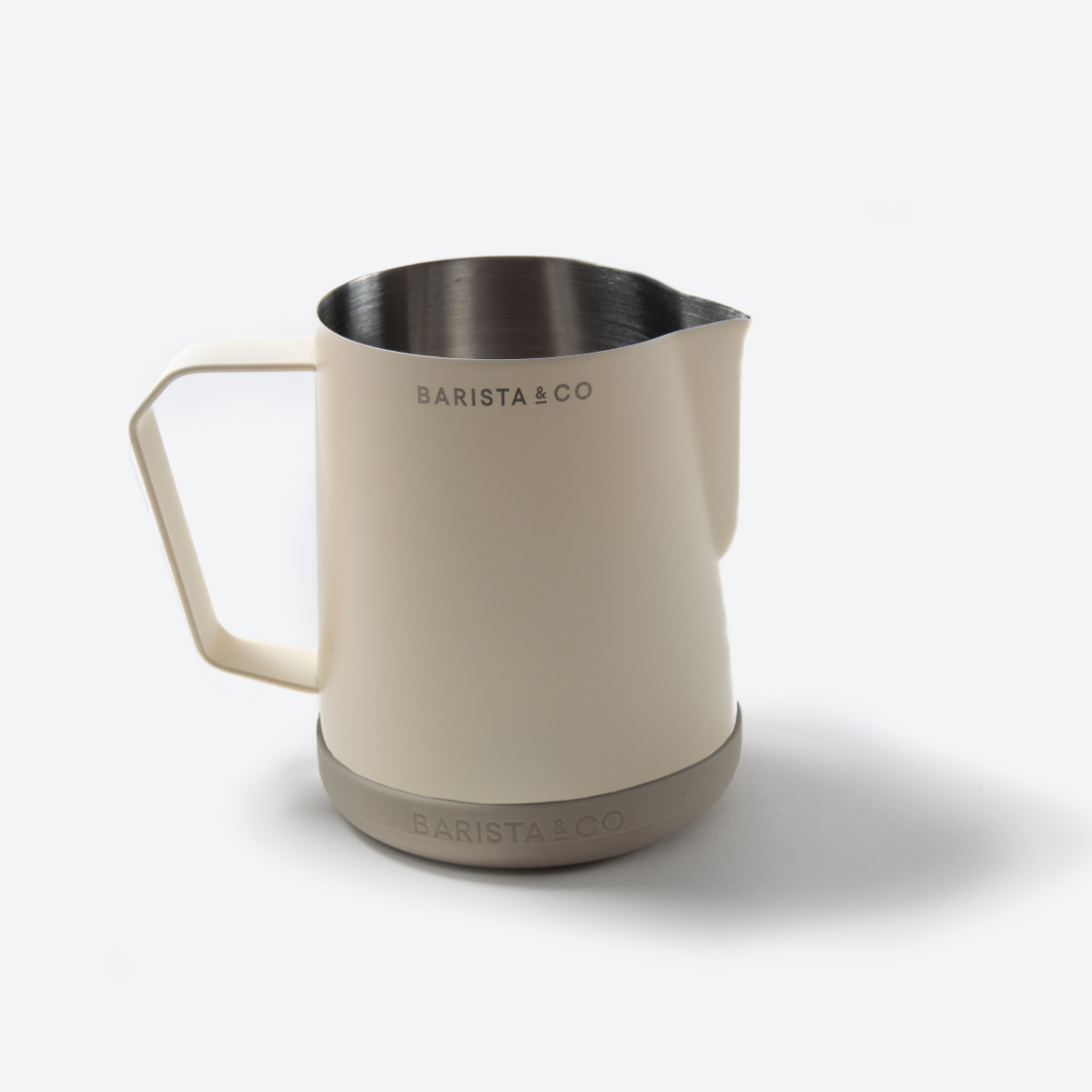A Barista & Co. stainless steel milk pitcher with a cream-colored handle and body, a brushed silver base, and black printed branding on the side, set against a clean white background.