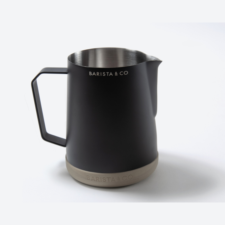 A matte black stainless steel milk pitcher with a handle, spout, and a light beige base, branded BARISTA & CO.