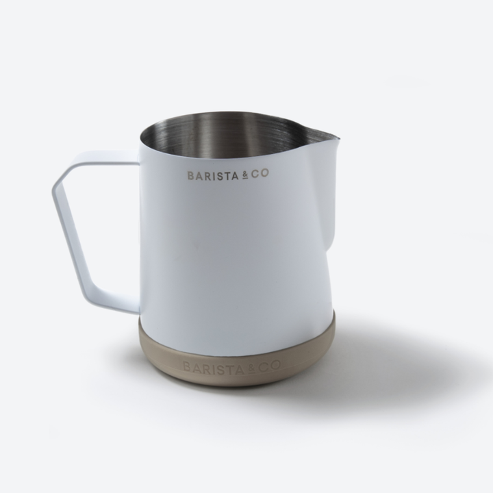A white Barista & Co stainless steel milk pitcher with a beige textured base and a white handle, shown on a light gray background.
