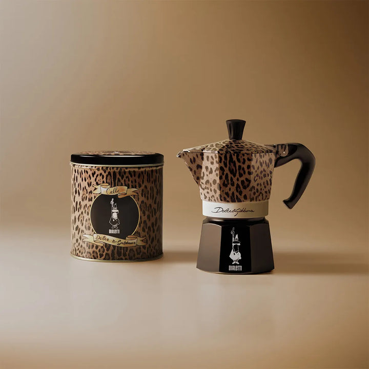A leopard-print moka pot coffee maker next to a matching cylindrical tin canister on a warm beige background. The canister features a black circular label with a white espresso cup illustration and the text 'Delizia' and 'Especialista'.