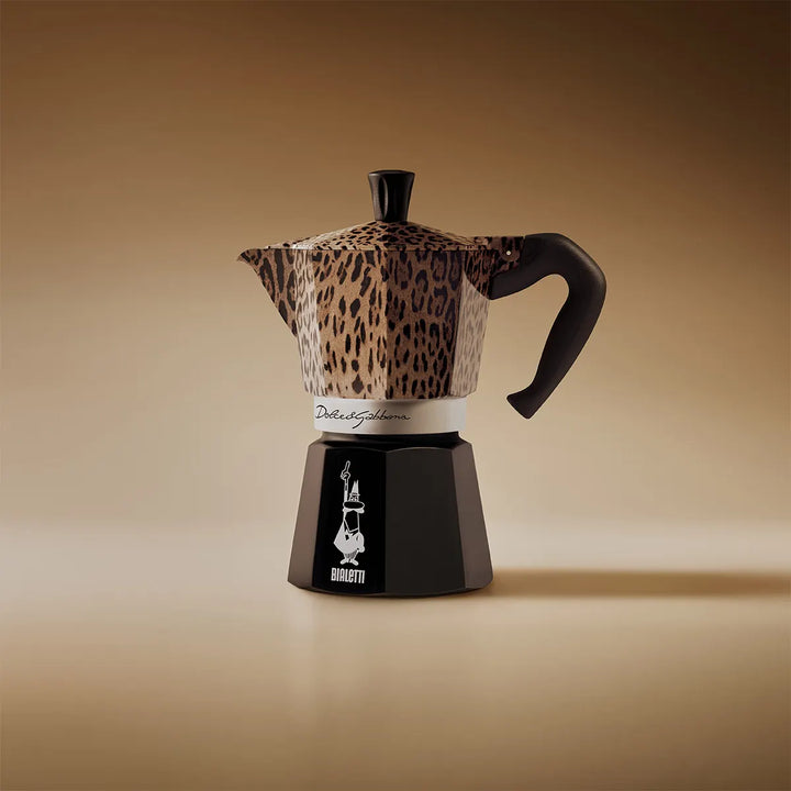 A Bialetti moka pot coffee maker with a leopard print pattern on the upper body and lid, paired with a black base and handle, set against a warm brown gradient background.